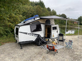 Enjoy the outdoors! This white Taxa Cricket rv rental trailer, with awning and chairs, is perfectly set up by a pond.