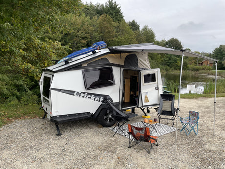 Enjoy the outdoors! This white Taxa Cricket rv rental trailer, with awning and chairs, is perfectly set up by a pond.