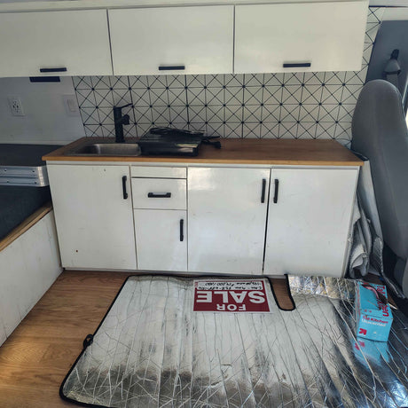A compact RV rental kitchen features white cabinets, wood counter, sink, black faucet, and a geometric backsplash. A "FOR SALE" sign is on a sunshade.