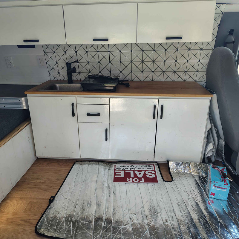 A compact RV rental kitchen features white cabinets, wood counter, sink, black faucet, and a geometric backsplash. A "FOR SALE" sign is on a sunshade. — photo 8