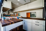 Cozy RV rental van interior features a bed with colorful quilt, kitchen sink, wooden counters, and white cabinets with windows.
