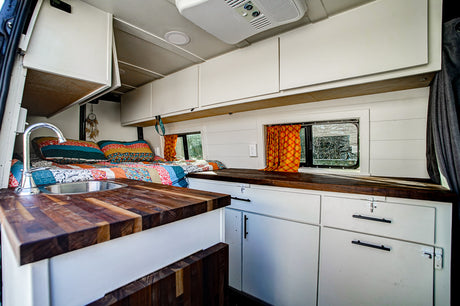 Cozy RV rental van interior features a bed with colorful quilt, kitchen sink, wooden counters, and white cabinets with windows.