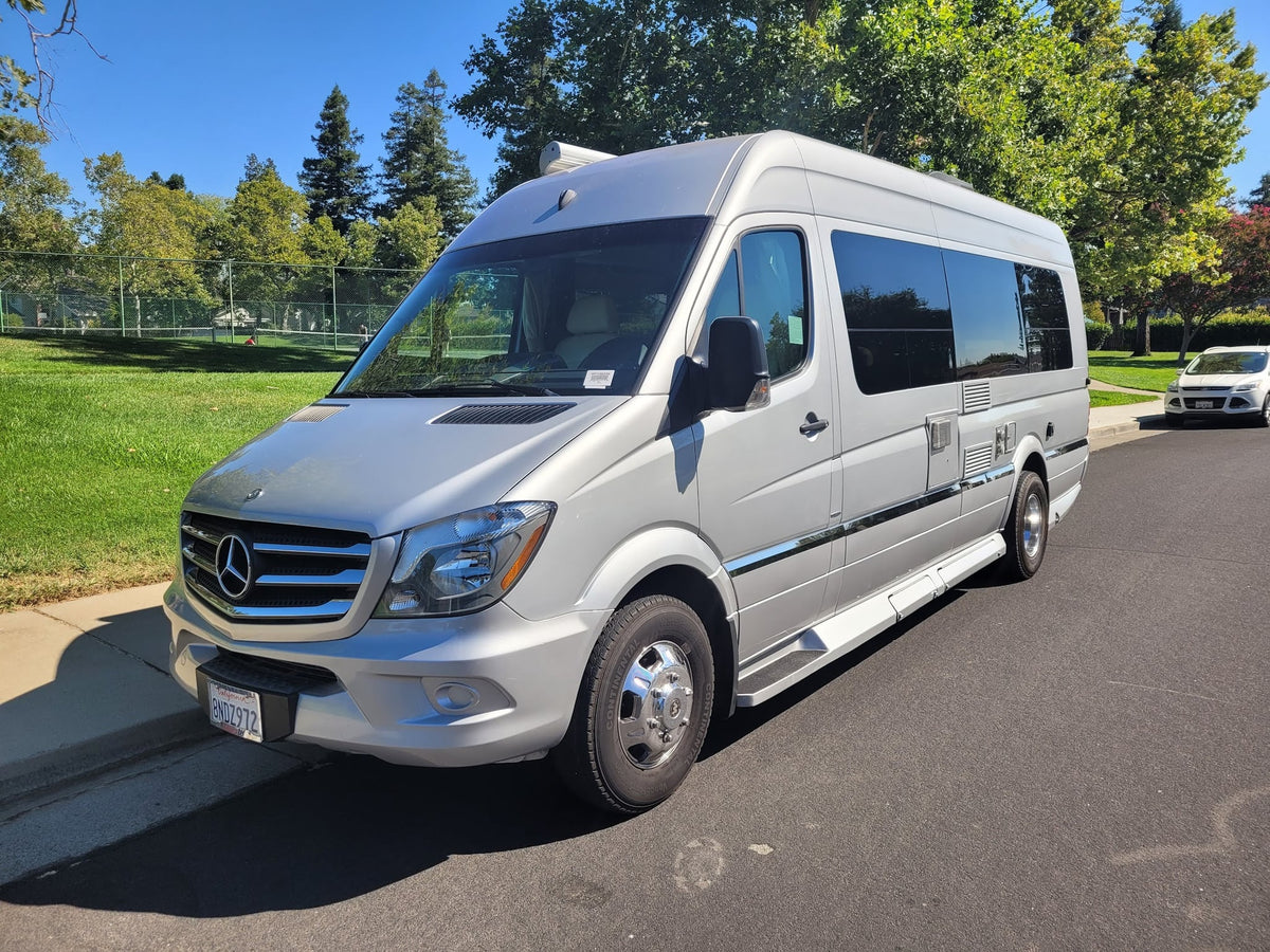 Explore in style with this silver Mercedes-Benz Sprinter RV rental. Parked curbside on a sunny day near a green park.|Front view of a silver Mercedes-Benz Sprinter RV rental van
