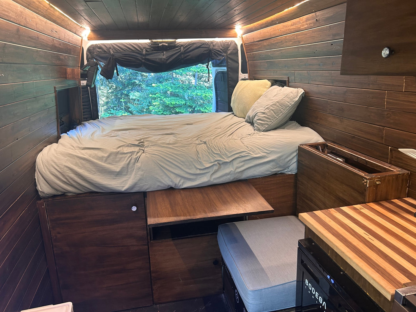 Cozy RV rental interior with a comfortable bed, warm wood paneling, and a serene nature view from the rear window. — photo 16