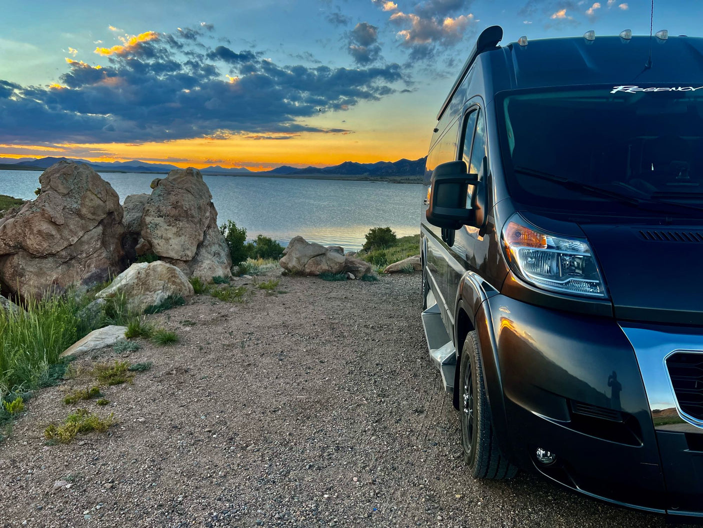 Dark RV rental parked on gravel, facing a lake and mountains under colorful sunset sky. Ideal for scenic road trips.