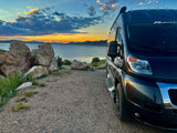 Dark RV rental parked on gravel, facing a lake and mountains under colorful sunset sky. Ideal for scenic road trips.