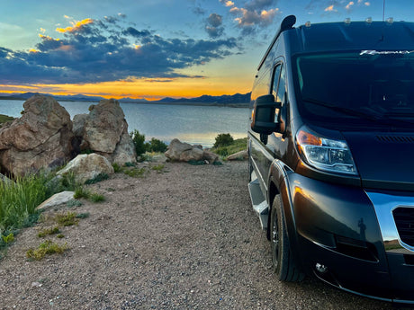 Dark RV rental parked on gravel, facing a lake and mountains under colorful sunset sky. Ideal for scenic road trips.