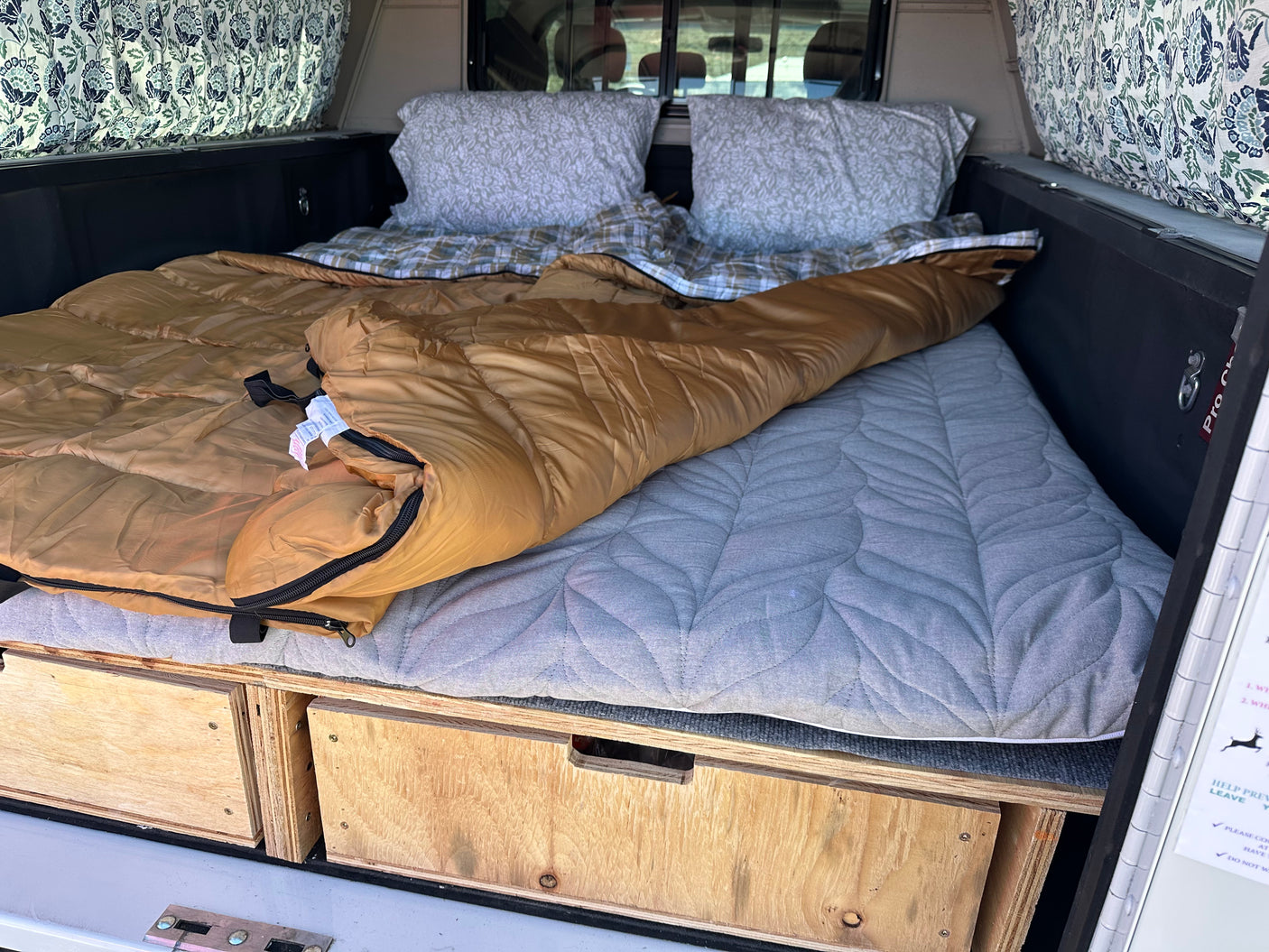 Cozy RV rental truck camper bed with a sleeping bag, pillows, and custom wooden storage drawers, ideal for mobile living.