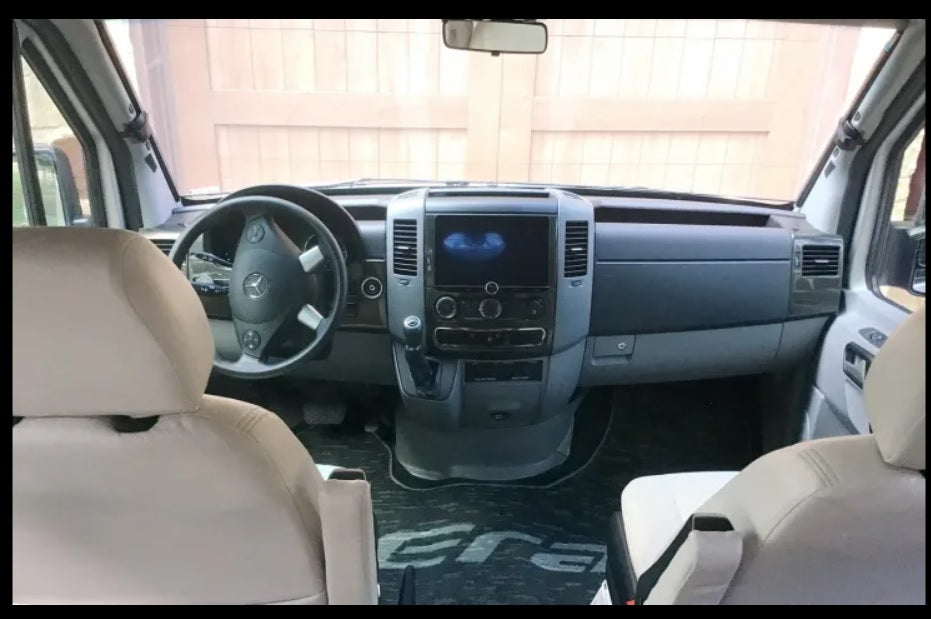 Front driving area of a Mercedes Sprinter Era RV rental. Dashboard, steering wheel, and light-colored front seats visible.