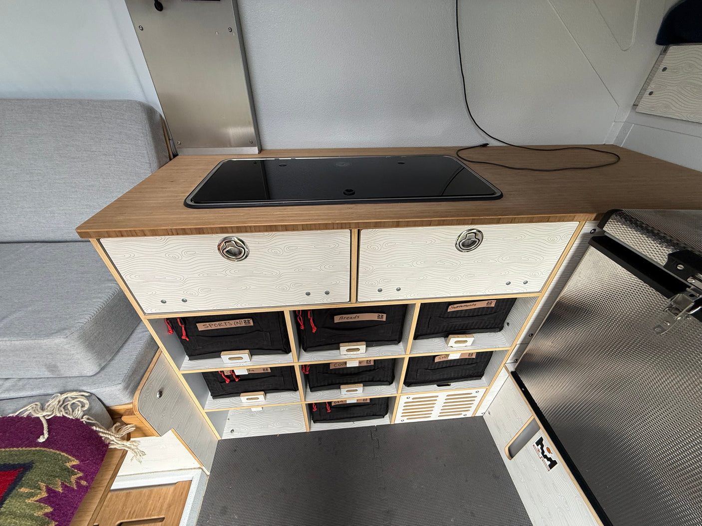 RV rental interior featuring a cooktop, custom labeled storage bins (Sports, Breads, Supplements), and a cozy seating area.