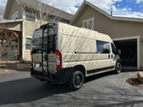 Explore this spacious RV rental! A beige campervan with a rear ladder is parked in a sunlit driveway by a modern home.