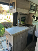 Inside an RV rental: a modern kitchenette with a bright counter, sink, induction cooktop, fridge, and overhead microwave.