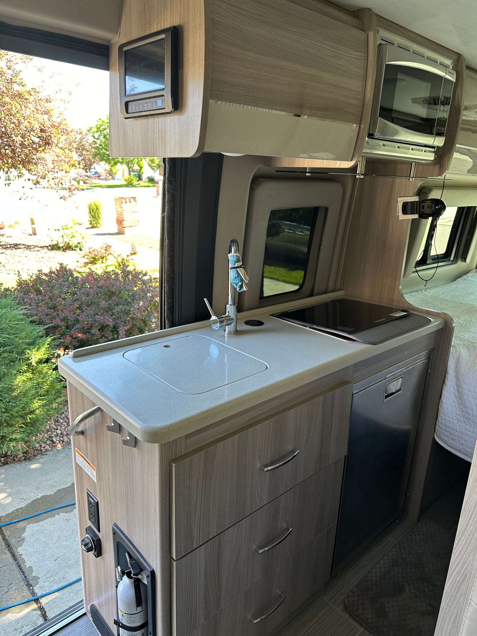 Inside an RV rental: a modern kitchenette with a bright counter, sink, induction cooktop, fridge, and overhead microwave.