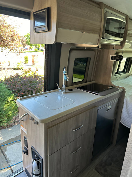 Inside an RV rental: a modern kitchenette with a bright counter, sink, induction cooktop, fridge, and overhead microwave.