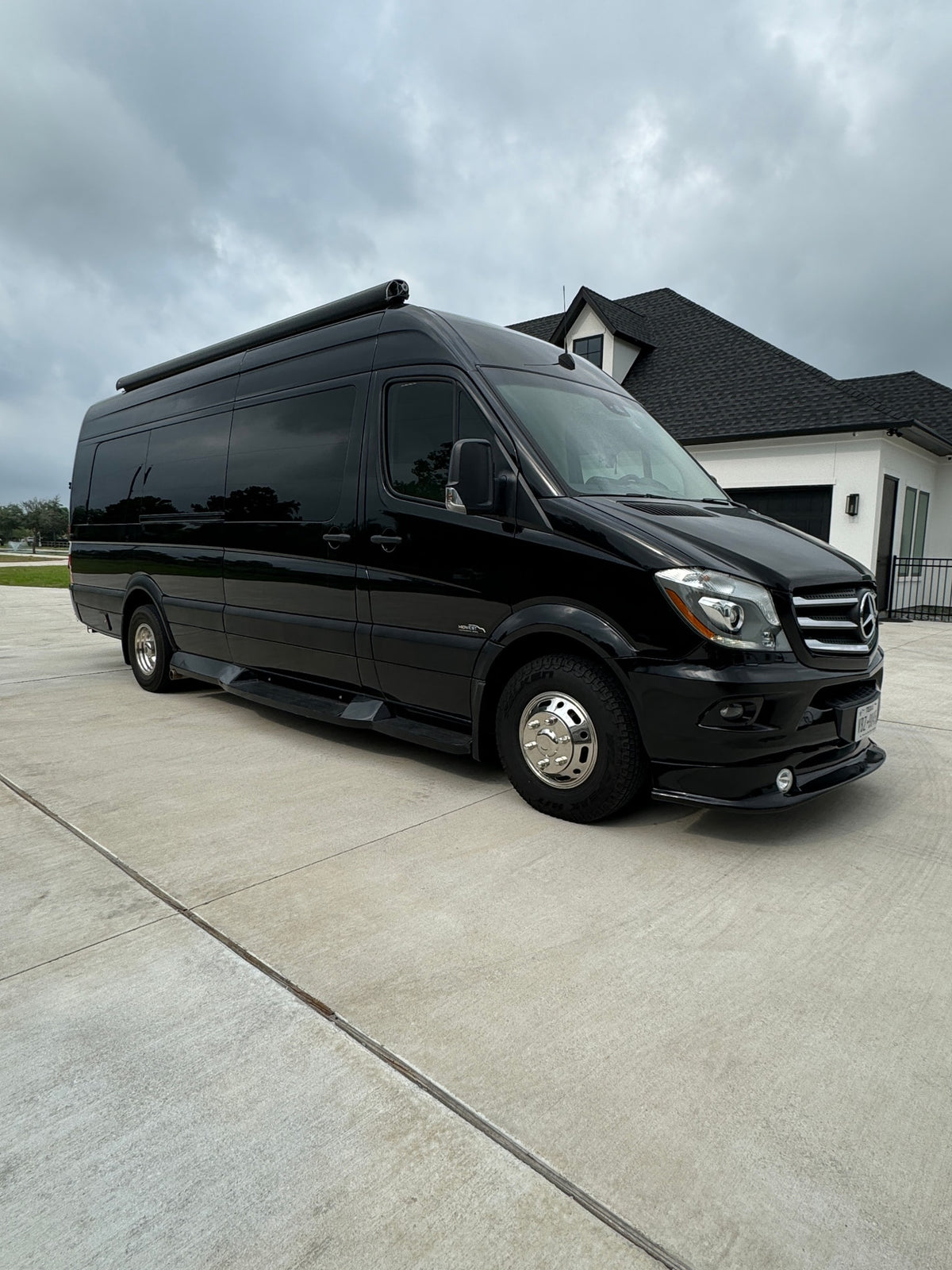 Black Mercedes-Benz Sprinter van, an ideal rv rental, parked on a concrete driveway by a house under cloudy skies.