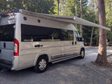 A silver Winnebago Travato RV rental is parked with its awning extended, offering shade at a wooded campsite.