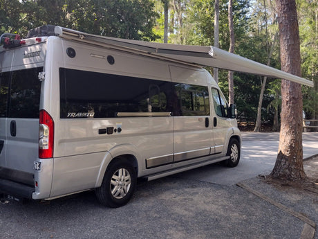 A silver Winnebago Travato RV rental is parked with its awning extended, offering shade at a wooded campsite.