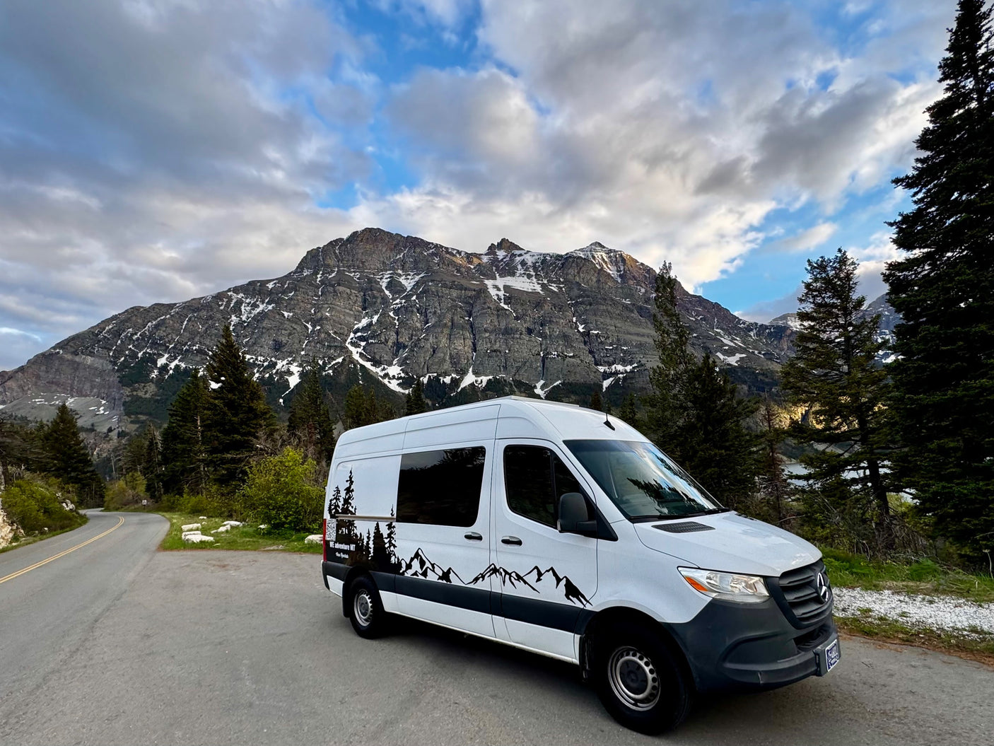 White RV rental van parked on a scenic mountain road with snow-capped peaks and pine trees under cloudy skies.