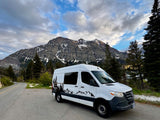 White RV rental van parked on a scenic mountain road with snow-capped peaks and pine trees under cloudy skies.