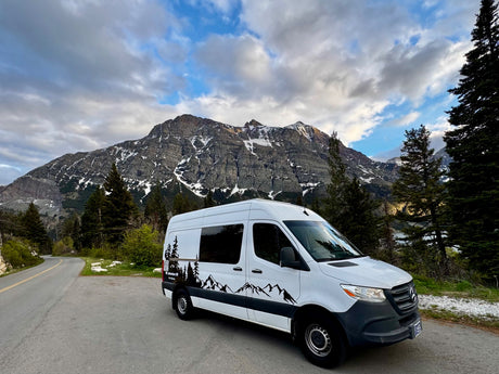 White RV rental van parked on a scenic mountain road with snow-capped peaks and pine trees under cloudy skies.
