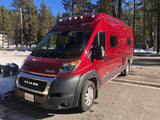 Red Winnebago Solis camper van (RV rental) among snowy pines, ready for winter adventure & cold-weather travel.