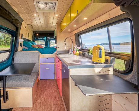 Bright camper van interior: wood walls, vibrant kitchen, cozy bed, dining area, and a large window view of the water, ideal for an RV rental.