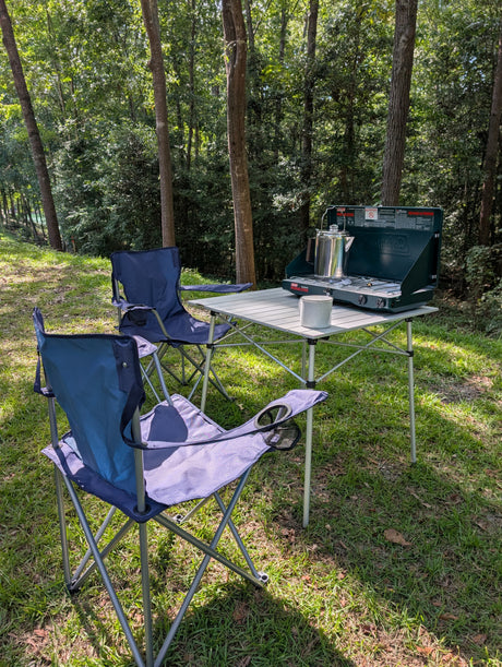 Perfect RV rental spot: chairs, table, stove, and coffee brewing in the peaceful woods.