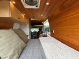 Cozy RV rental interior with a comfy bed, warm wood paneling, and a compact kitchen. View extends to the driver's cab.