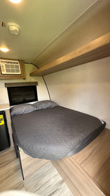 Cozy RV rental bedroom features a gray bed, AC unit, mini-fridge, and an overhead shelf, with a window and smoke detector.