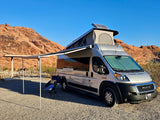 Silver RV rental with pop-top and extended awning at a desert campground with red rock scenery.