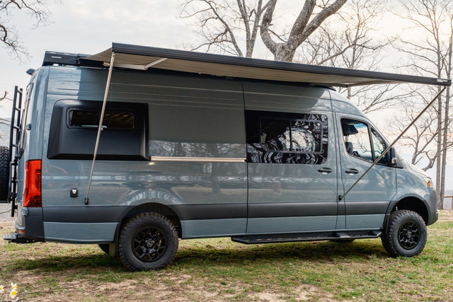 Gray Sprinter camper van with an extended awning, off-road tires, and rear ladder, ideal for your next RV rental adventure.