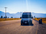 A white camper van (RV rental) rests on a sunlit rural road, with golden fields, fences, and blue mountains in the distance. Ideal for scenic road trips.