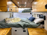 Inside an rv rental camper van: a cozy bed with green blankets, wooden interiors, and a view of a rocky riverbed and mountains.