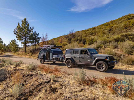 Grey Jeep tows a rugged off-road RV rental trailer on a dirt road. Perfect for exploring hills & nature under a blue sky!