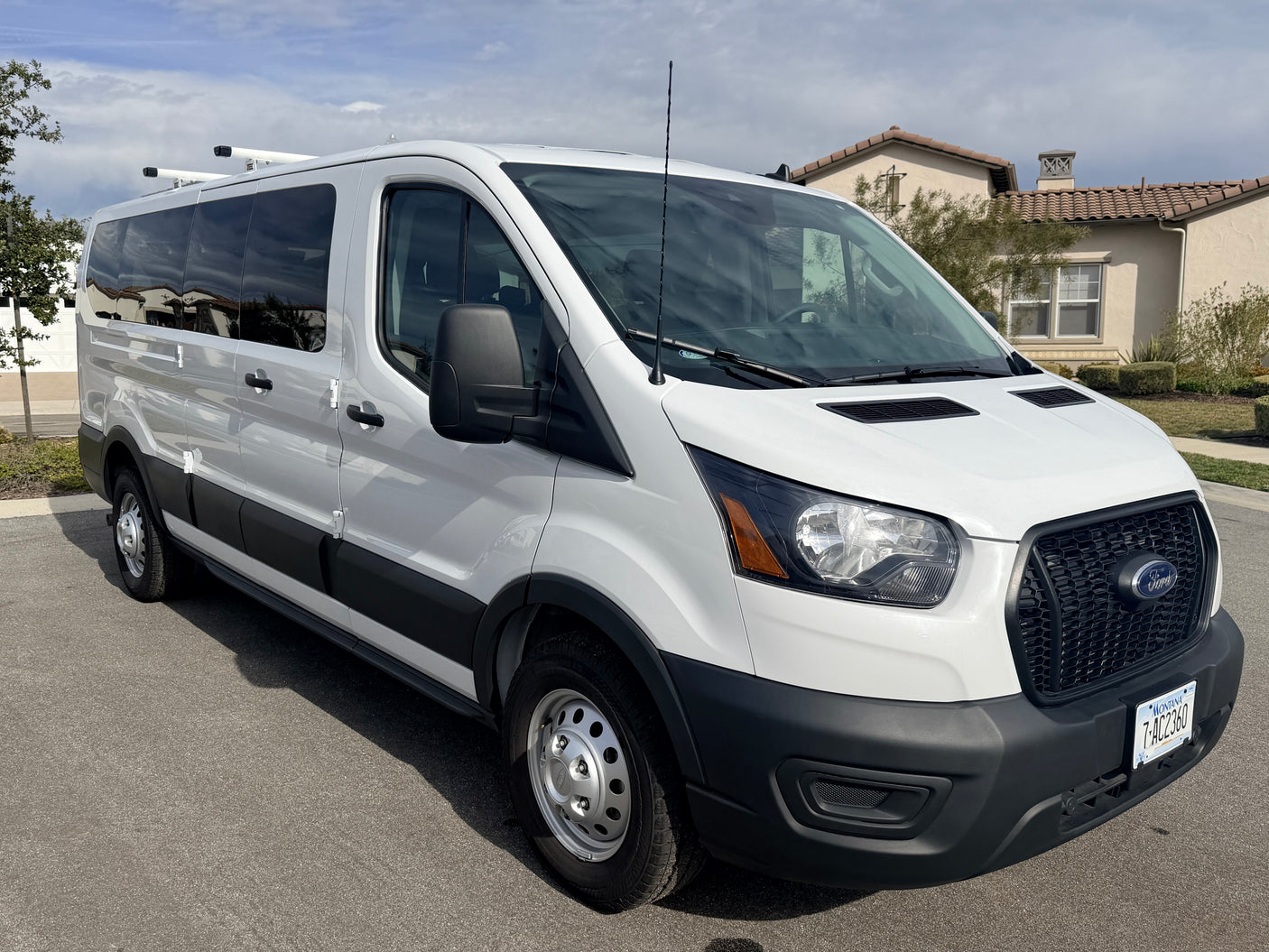A white Ford Transit van with roof racks, perfect for an RV rental adventure. Parked outdoors on asphalt with houses in the background.
