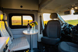 Cozy RV rental interior at sunset: table with wine & flowers, comfy seats, driver's area visible.