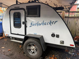 A compact white Bushwhacker teardrop RV rental, featuring off-road tires, is shown parked outdoors on a gravel surface.