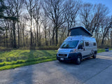 Light gray camper van with pop-top parked on asphalt next to a sunny forest. Perfect for an rv rental trip.