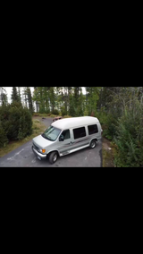 An RV rental silver van awaits adventure at a tranquil forest campsite with a lake in the background.