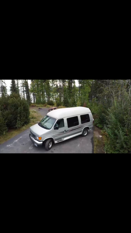 An RV rental silver van awaits adventure at a tranquil forest campsite with a lake in the background.