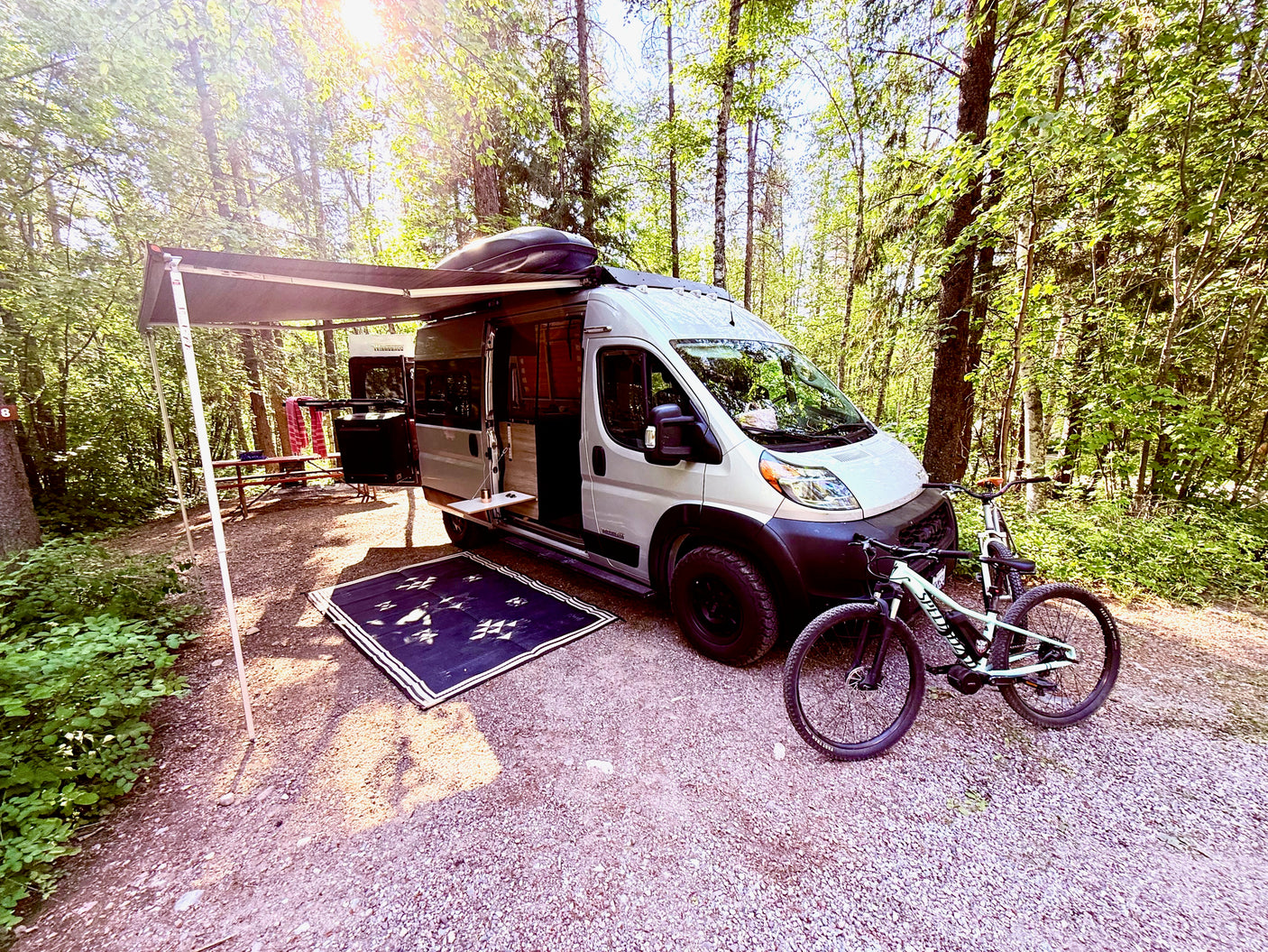 An RV rental camper van parked at a sunny forest campsite, with an extended awning, outdoor mat, and bicycles.