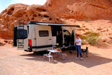 RV rental camper van parked in a stunning red rock desert. Woman, outdoor kitchen, & chairs set up for a scenic camp.