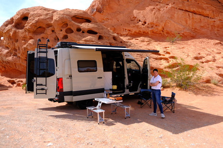 RV rental camper van parked in a stunning red rock desert. Woman, outdoor kitchen, & chairs set up for a scenic camp.
