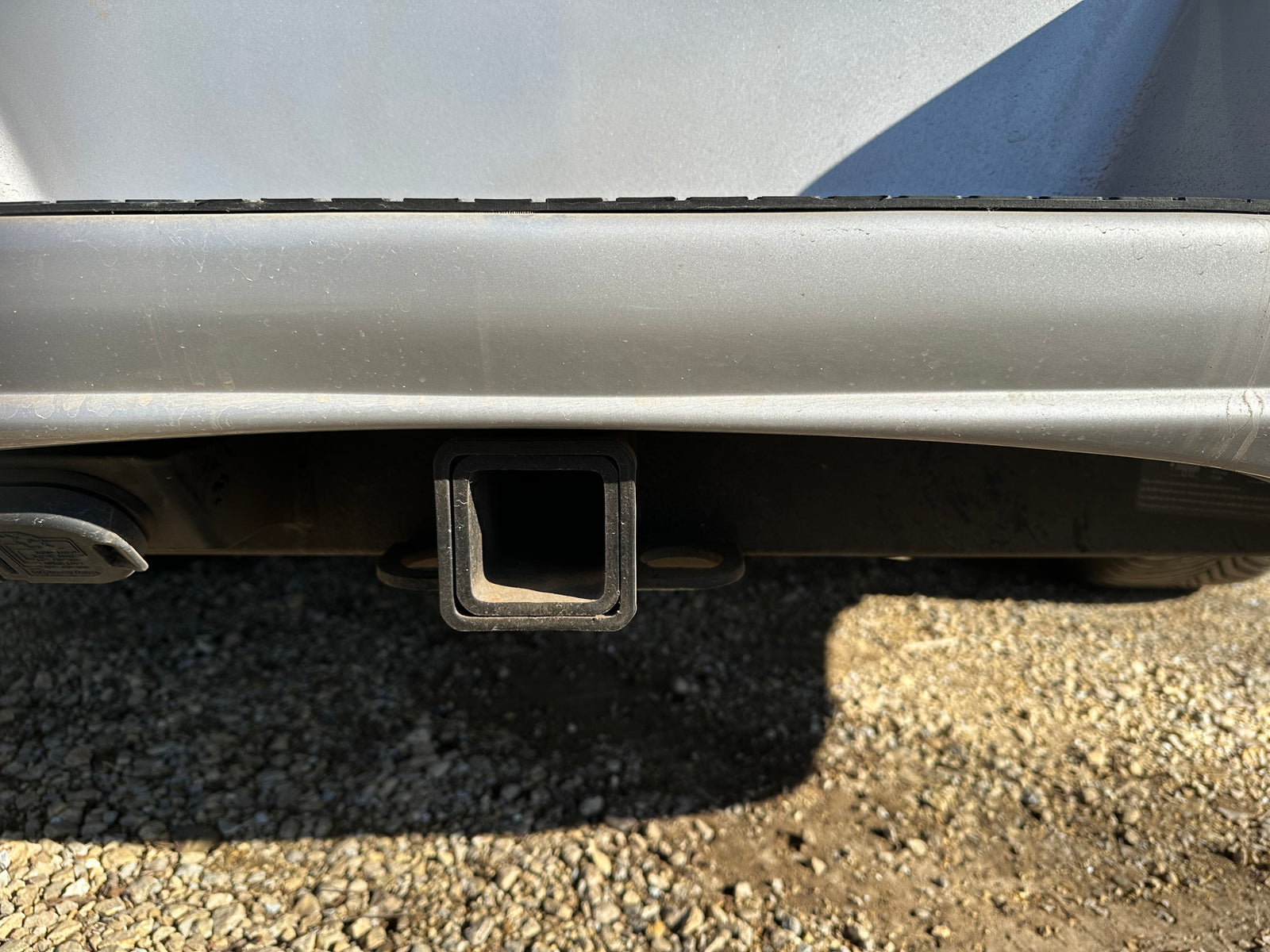 Silver RV rental's rear with trailer hitch receiver and electrical connection for towing, on gravel.