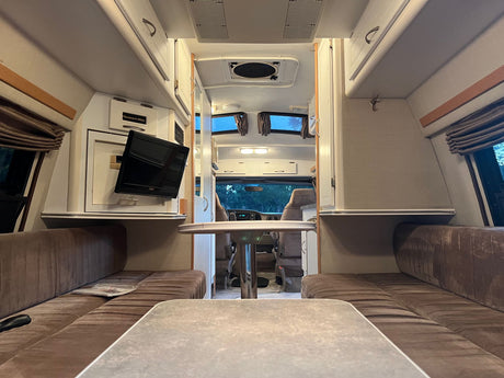 Longitudinal view of a cozy RV rental interior with seating, dual tables, and TV, facing the driver's cab.