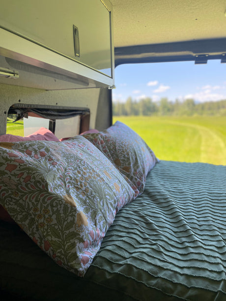Cozy RV rental bed with patterned pillows, a textured green comforter, and a stunning view of a bright green field.