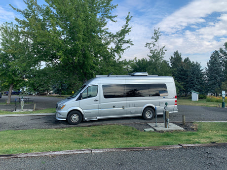 A silver Airstream Interstate RV rental parked on gravel at a leafy campsite with utility hookups. Perfect for travel!