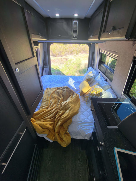RV rental: a cozy interior featuring a bed with yellow accents, compact kitchen, and scenic nature view from the rear.