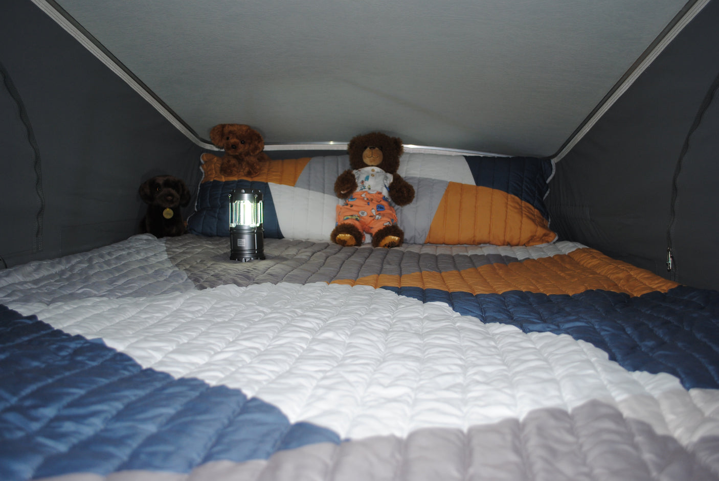 Inside an RV rental: a cozy bed with a colorful quilt, a glowing lantern, and three stuffed animal companions.