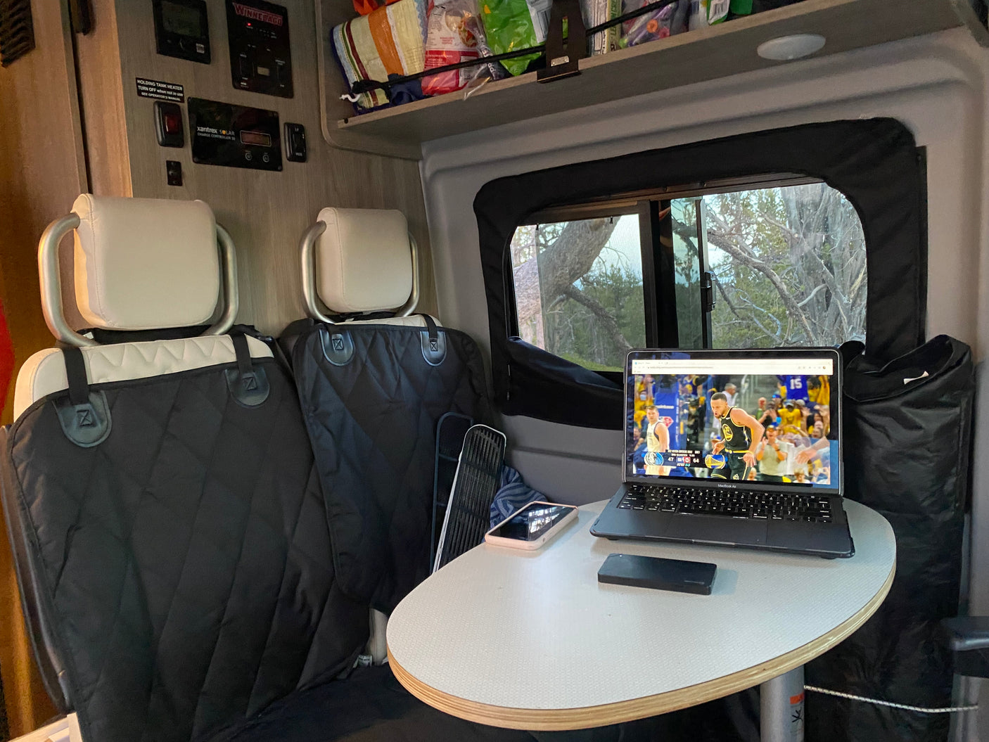 An RV rental provides a mobile office and entertainment hub, with a laptop showing a basketball game and scenic window views.